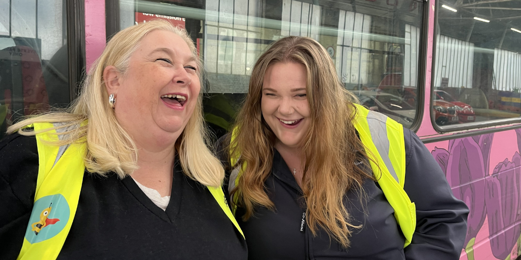 two women wearing hi-vis jackets laughing in front of a bus